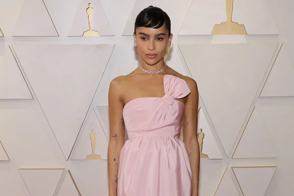 A atriz Zoë Kravitz no tapete vermelho do Oscar 2022. Ela é uma mulher negra, com cabelo presto e uma franja curta, e usa um vestido rosa claro da marca Yves Saint Laurent