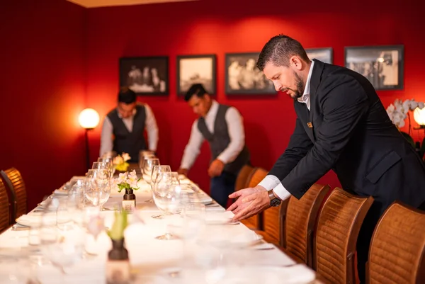 Duas pessoas, um homem e uma mulher, servem vinho e conversam sobre a mesa, iluminada por velas. Ao fundo, prateleiras exibem garrafas de rótulos premium.