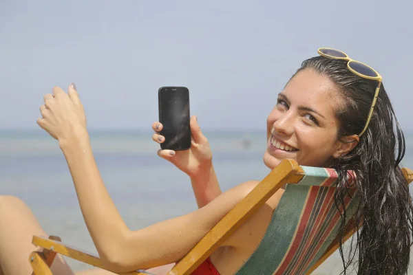 Foto colorida de mulher sentada em cadeira de praia e segurando celular - Metrópoles