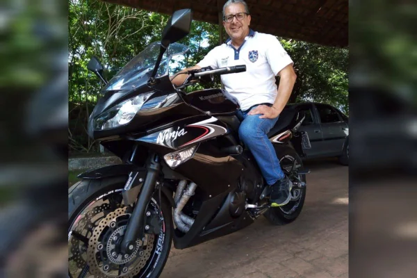 Homem branco de camiseta branca e calça jeans sorri montado em uma moto - Metrópoles