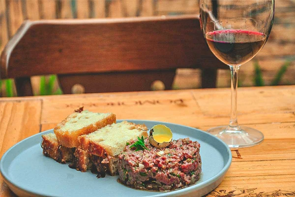 Prato com steak tartare e torradas e taça de vinho, tudo sobre uma mesa de madeira
