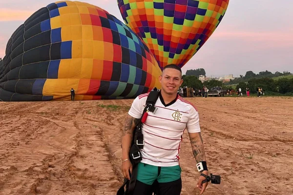 Homem forte, de cabelo raspado, vestindo camiseta do Flamengo segura câmera em uma das mãos; ao fundo, dois balões coloridos
