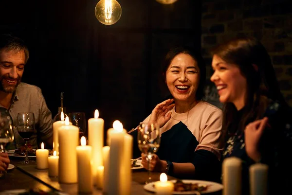 Foto colorida de duas mulheres e um homem sentados à mesa com velas acesas em um jantar - Metrópoles