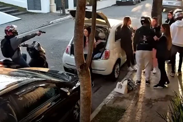 Imagem colorida em que três homens armados rendem cinco mulheres em frente a um carro
