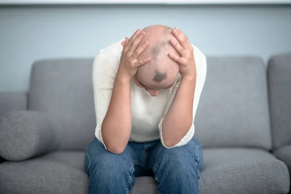 Pessoa sentada em um sofá olhando para baixo. Ela tem a cabeça raspada alopecia - Metrópoles