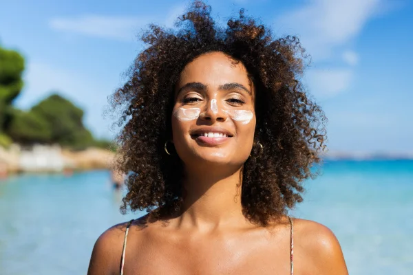 Foto colorida de mulher em praia e com protetor solar no rosto - Metrópoles