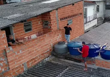 Imagem aérea feita por drone de dois homens, um de camiseta preta e outro de vermelho, em cima de um telhado; outro homem de camiseta preta está em uma sacada, apontando arma - Metrópoles