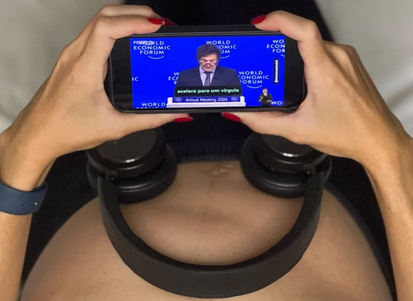 Fotografia colorida mostra barriga de Marina Helena, que está grávida, com fones de ouvido posicionados sobre ela e o discurso de Javier Milei transmitido em tela de celular - Metrópoles