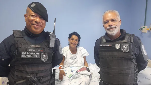 Imagem colorida de uma mulher segurando um bebê e dois policiais sorrindo - Metrópoles