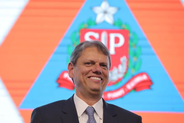 Imagem colorida mostra governador de São Paulo, Tarcísio de Freitas, sorrindo com os dentes à mostra. Ele é um homem branco com cabelos grisalhos e veste blazer azul marinho, camisa branca e gravata azul. Ao fundo, na imagem, aparece a bandeira de São Paulo - Metrópoles