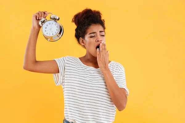 Foto mostra mulher negra bocejando e segurando um despertador. Soneca - Metrópoles