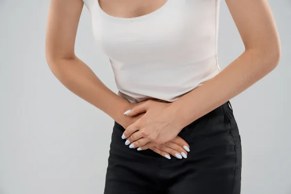 Close-up de uma mulher caucasiana vestindo um conjunto casual, inclinada e posando sobre um fundo cinza isolado, enquanto toca a parte inferior do abdômen devido a uma dor. Enquadramento frontal de uma mulher de cabelos castanhos sofrendo de dor no estômago. Metrópoles