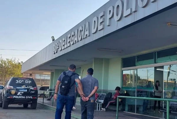 foto colorida de dois homens de costas e um algemado diante de delegacia
