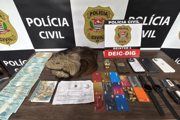 Imagem colorida de uma mesa da Polícia Civil com diversos itens apreendidos na operação. Cartões bancários, dinheiro em espécie, aparelhos eletrônicos e perucas foram encontrados - Metrópoles