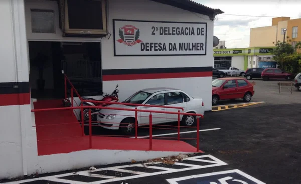 Entrada de delegacia, um edifício cor branca com faixas em preto e vermelho; há um carro e uma moto estacionados - Metrópoles