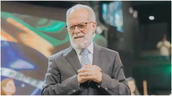 Márcio Valadão, pai do também pastor André Valadão durante culto. Ele usa terno, óculos e microfone e fala para uma plateia com bandeira do Brasil em telão atrás - Metrópoles