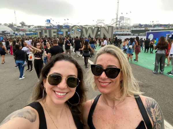 Imagem colorida mostra duas mulheres vestidas de camisetas regadas pretas, de óculos escuros, sorrindo enquanto uma multidão de pessoas anda atrás delas no festival The Town - Metrópoles