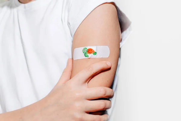Foto colorida em close do braço de uma criança com a camiseta branca e um band-aid colado no braço com um desenho de tartaruga - Metrópoles