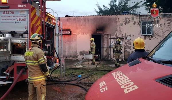 Quatro bombeiros do Corpo de Bombeiros do Distrito Federal em meio à incêndio. Ao fundo, casa suja pela fuligem, ambulância no canto direito e caminhão de bombeiros no canto esquerdo.