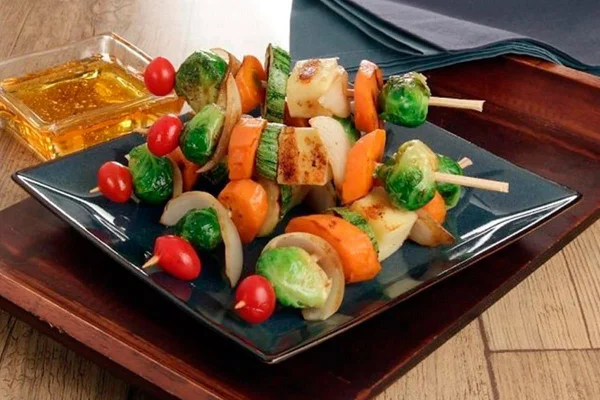 Espetinho de legumes com queijo coalho em travessa quadrada. Ao lado, pote de vidro com molho amarelo - Metrópoles