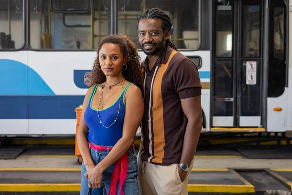 foto colorida de uma mulher e um homem negros posando juntos com um ônibus ao fundo - metrópoles