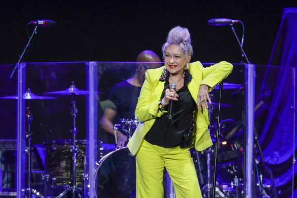 Em palco, cantora Cyndi Lauper segura microfone. Ela usa blusa preta com blazer e calça amarelos - Metrópoles