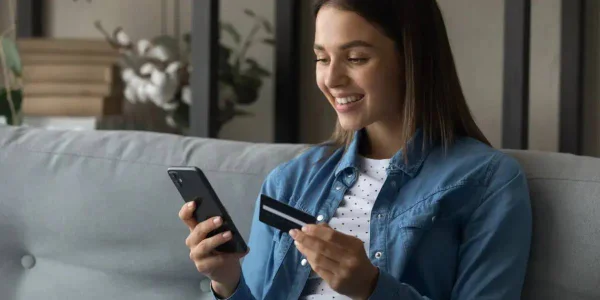 Mulher sorrindo e sentada em sofá segura cartão de crédito e celular nas mãos - Metrópoles