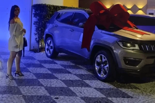 Imagem colorida de uma mulher de vestido branco ao lado de um carro cinza com um laço vermelho em cima - Metrópoles