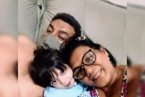 Homem e mulher abraçando criança no meio, deitados em sofá cinza