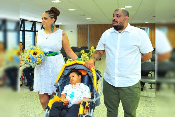 Imagem colorida de um homem e uma mulher vestidos de branco. Em meio aos dois, está um carrinho de bebê com um garoto de aproximadamente 3 anos dentro. A mulher, à direita, está segurando um buquê nas mãos