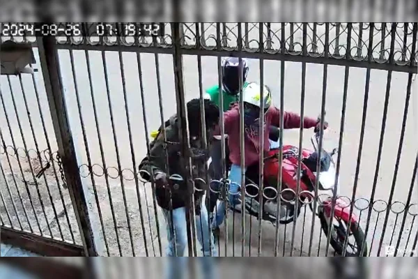Imagem de dois homens em uma moto vermelha abordando um adolescente de casaco preto e calça azul em frente a um portão de grades finas