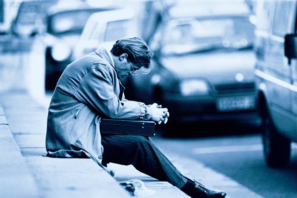 Imagem em preto e branco de homem sentado na calçada, de cabeça baixa, com os braços sobre uma pasta, com carros passando ao fundo - Metrópoles