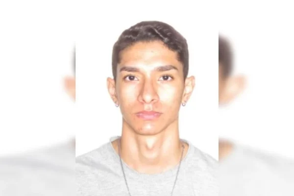 Imagem colorida da foto 3x4 de um jovem branco, magro, de cabelo curto, corrente prata no pescoço e camiseta cinza