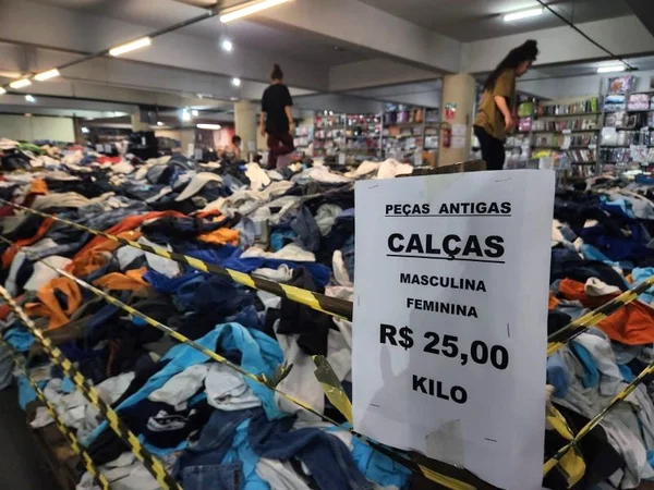 imagem colorida mostra duas pessoas em cima de pilha de roupas. metrópoles