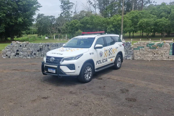 Imagem colorida mostra carro branco com a palavra Polícia escrita na lateral. Ao fundo está um paredão formado por pacotes em formato de tijolos que ocupam toda a extensão horizontal da foto - Metróopoles