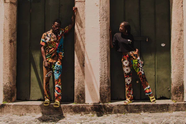 Duas pessoas apoiadas em uma pilastra. A dupla usa roupas coloridas de verão / Metrópoles