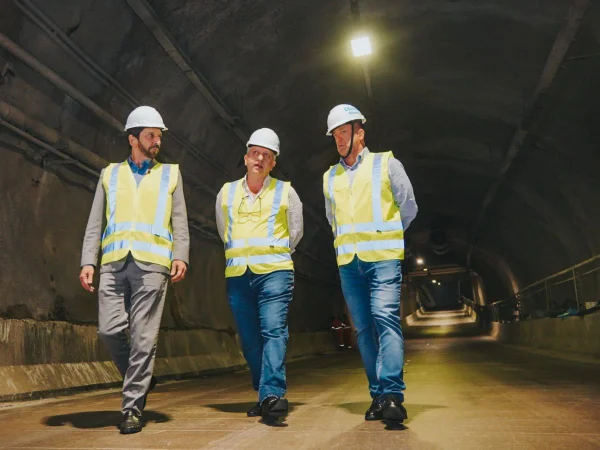 Imagem colorida mostra o prefeito Ricardo Nunes e mais dois homens andando em um túnel viário vazio. Eles vestem capacete branco e colete fluorescente - Metrópoles