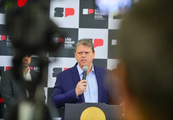 Imagem colorida mostra Tarcísio de Freitas, homem branco, grisalho, de terno azul marinho e camisa azul clara, entre câmeras de TV, dando uma entrevista - Metrópoles