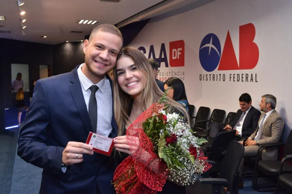 Homem de cabelo curto e terno abraça mulher de preto. Ele segura buquê de flores