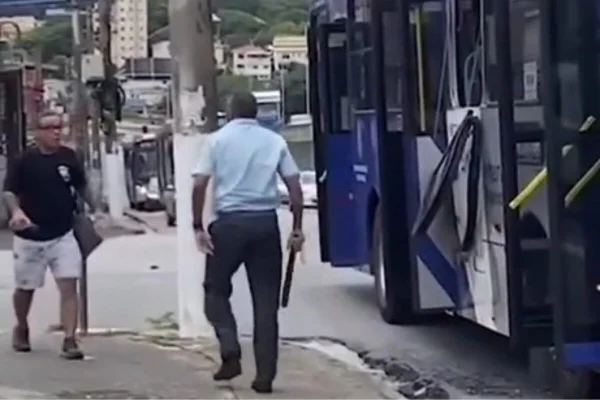 Foto colorida mostra motorista de ônibus segurando pedaço de pau caminhando em direção a passageiro, em uma calçada, ao lado de um ônibus da EMTU parado na rua - Metrópoles