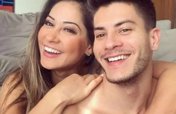 Na imagem colorida, duas pessoas estão posicionadas no centro. A mulher está do lado direito e abraça o homem que está do lado esquerdo