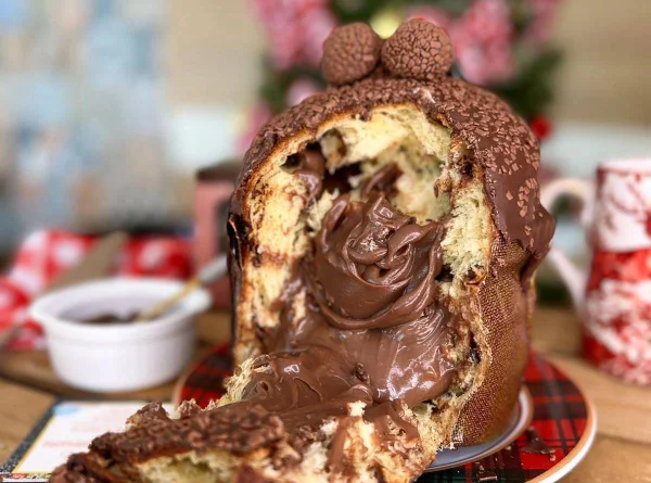 Na foto temos uma decoração de natal desfocada ao fundo e em primeiro plano um chocotone cortado com brigadeiro escorrendo. No topo dele, temos dois brigadeiros enrolados