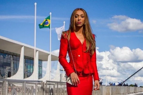 imagem colorida mostra a deputada federal erika hilton, uma mulher trans negra com cabelos loiros e lisos usando terno de cor vermelha, em frente ao palácio do planalto em brasília