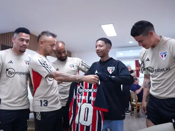 Foto colorida mostra garoto no centro, de casaco preto do São Paulo, recebendo uma camisa das mãos do jogador Rafinha, que está ao seu lado. Os atletas James Rodrigues, Lucas Moura e Luciano também aparecem.