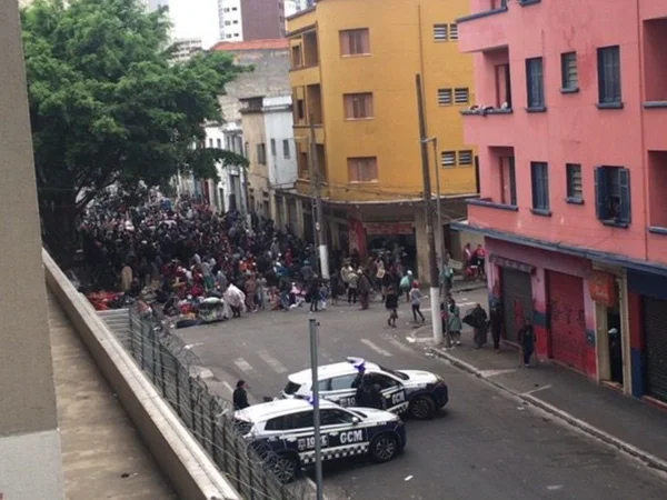 Imagem colorida mostra cruzamento de ruas da Cracolândia, no centro de SP, com uma multidão de usuários de crack ao fundo e dois veículos policiais perto - Metrópoles