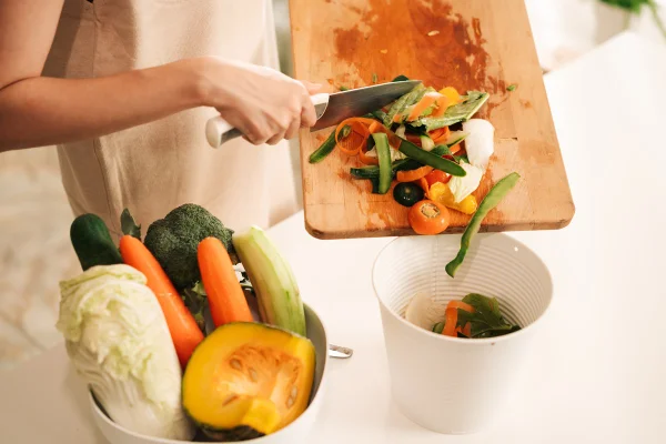 Foto colorida - Dois potes, um com legumes, como cenoura e abobora, e outro com cascas. Uma pessoa está jogando as cascas de uma tábua em frente do segundo pote