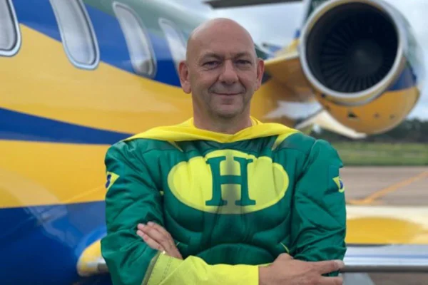 Fotografia colorida. Luciano posa com os braços cruzados e fantasiado de herói em frente a avião