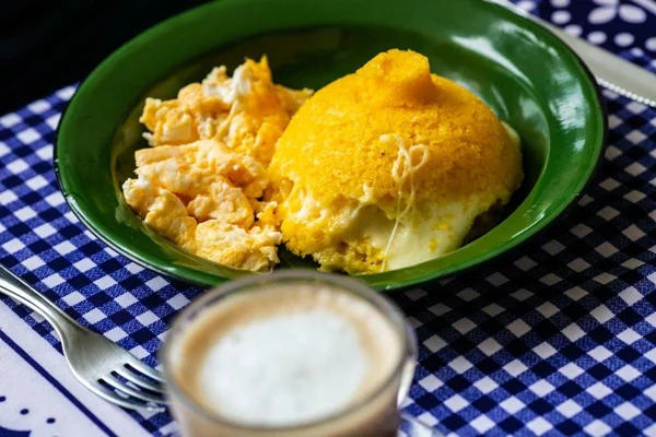 Prato com cuscuz com queijo e ovo e um copo de café com leite ao lado