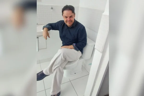 Imagem colorida mostra homem branco de cabelos curtos escuros. Ele veste calça branca, sapatos pretos e camisa de mangas compridas azul marinho. O homem está dentro de um banheiro com azulejos brancos, ele está sentado com pernas cruzadas em cima de uma privada com a tampa fechada - Metrópoles