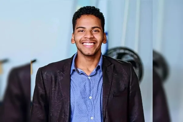 Imagem colorida de um homem negro, de camiseta azul e jaqueta preta, sorrindo